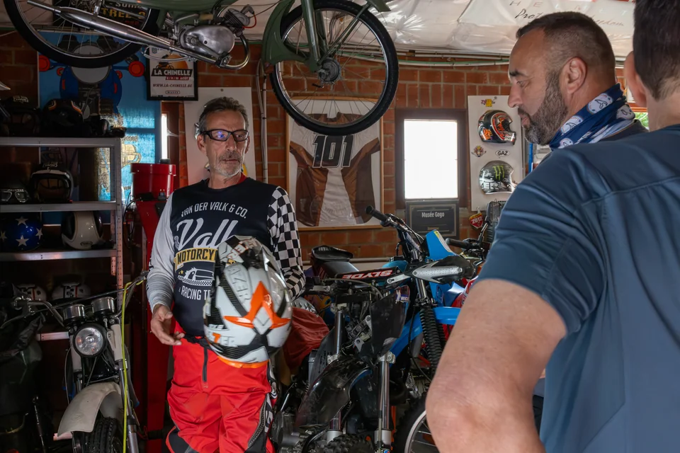 Olivier et un visiteur se tiennent dans un garage rempli de motos et de matériel.