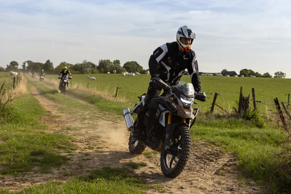 Un pilote approche sur un chemin de terre entre des champs, suivi d’autres motos.