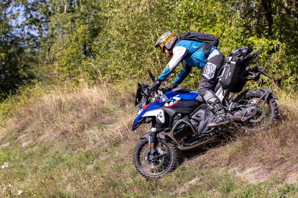 Un pilote sur une BMW trail négocie une pente herbeuse bordée d’arbres.