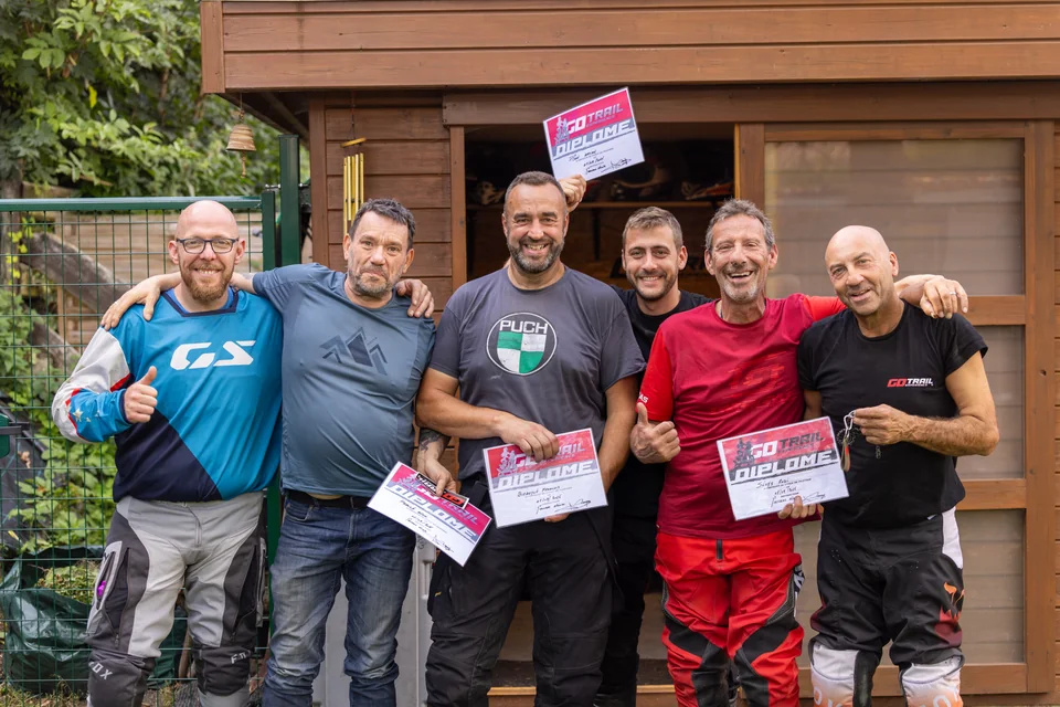 Six hommes posent ensemble devant un abri en bois, plusieurs avec leur diplôme.